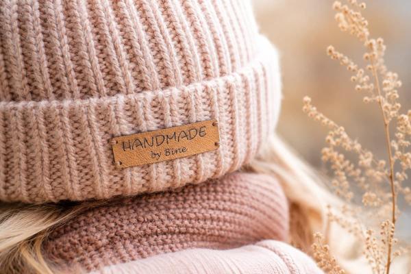 Rectangular tan faux leather label reading "Handmade by Bine" on a pink knit beanie.